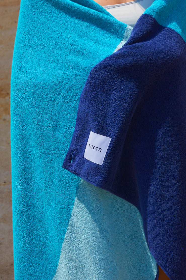 Fille embrassant une serviette de plage Tucca "Swell". Des couleurs bleu clair, bleu vert et bleu foncé en gros blocs composant un beau dessin. Grande serviette de plage qui ne se laisse pas souffler par le vent. Texture épaisse et douce car elle est faite de 100% coton.