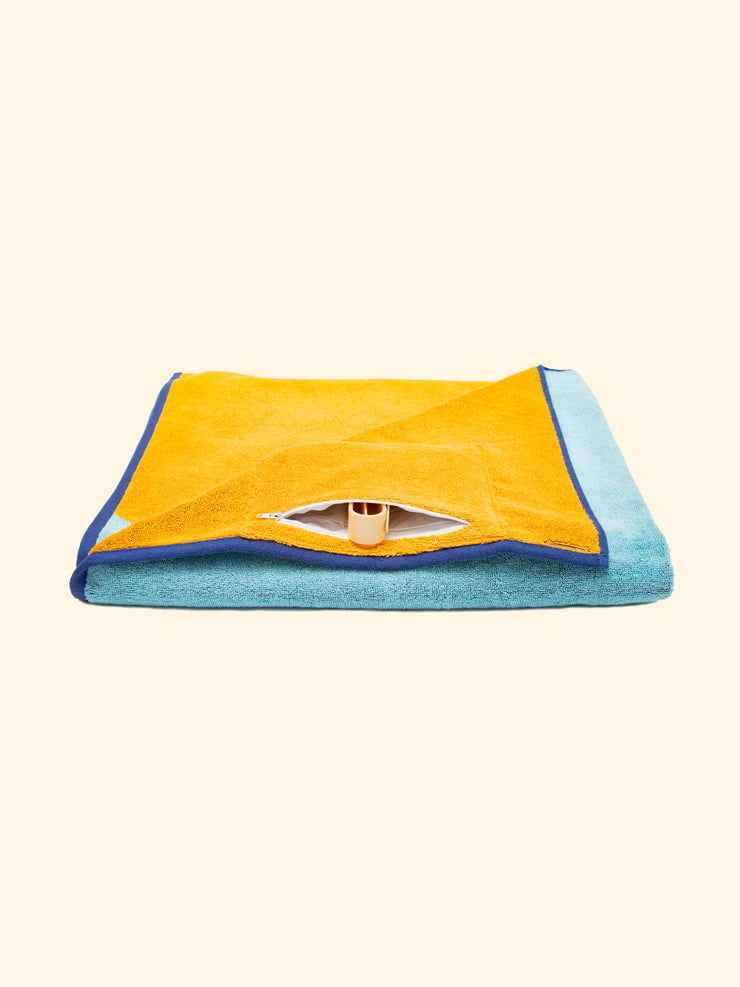 Modèle "Dune" de serviette de plage Tucca pliée tout en montrant sa poche cachée imperméable à fermeture éclair, qui peut être utilisée pour ranger votre téléphone ou d'autres objets ainsi que pour garder vos épingles Tucca comme le montrent les photos. Celles qui peuvent être utilisées pour fixer votre serviette de plage sur le sable.