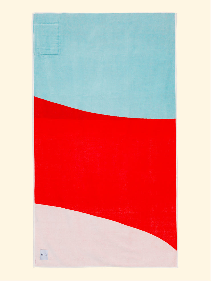 Serviette de plage Tucca "Berry" étendue. Couleurs bleu clair, rouge et blanc en gros blocs composant un beau dessin. Grande serviette de plage qui ne se laisse pas souffler par le vent. Texture super douce car elle est faite de coton 100% biologique.