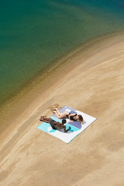 Two beach towels connected without sand in between and creating a bigger common space in the beach. Couple enjoying the sun in the beach lying on the Florida and Mayeri styles, premium beach towels, extra light and made of 100% organic cotton.