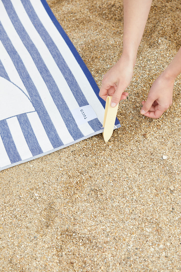 Photo montrant comment fixer votre serviette de plage dans le sable afin qu'elle ne soit pas emportée par le vent. Insertion de l'épingle Tucca dans le coin de la serviette de plage premium light style Floride. Fabriquée en 100% coton biologique.