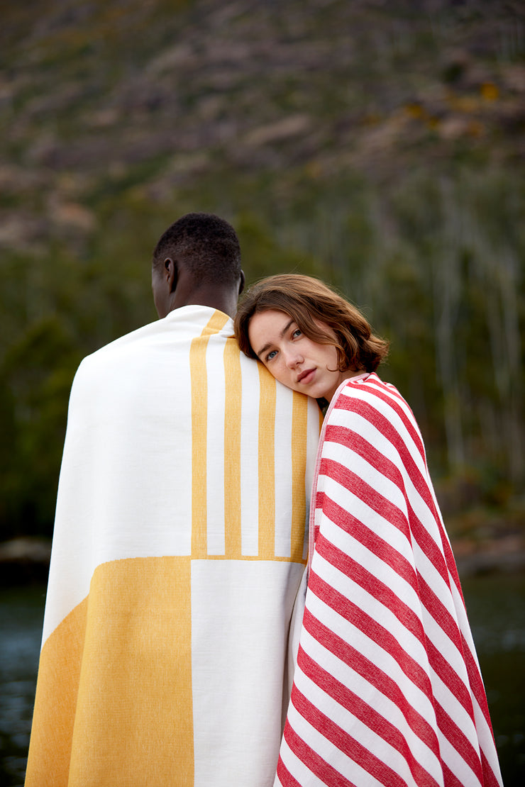 Un couple se séchant avec les serviettes de plage connectables Danai et Crassa. Elle s'appuie sur son épaule, tous deux recouverts de la serviette de plage Tucca light premiums en coton biologique.