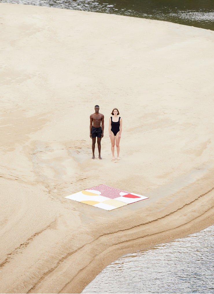 Deux serviettes de plage reliées l'une à l'autre. Les photos montrent les designs connectables des serviettes de plage Danai et Crassha premium ligh. Un couple profite de la plage ensemble avec leurs serviettes de plage reliées ensemble.